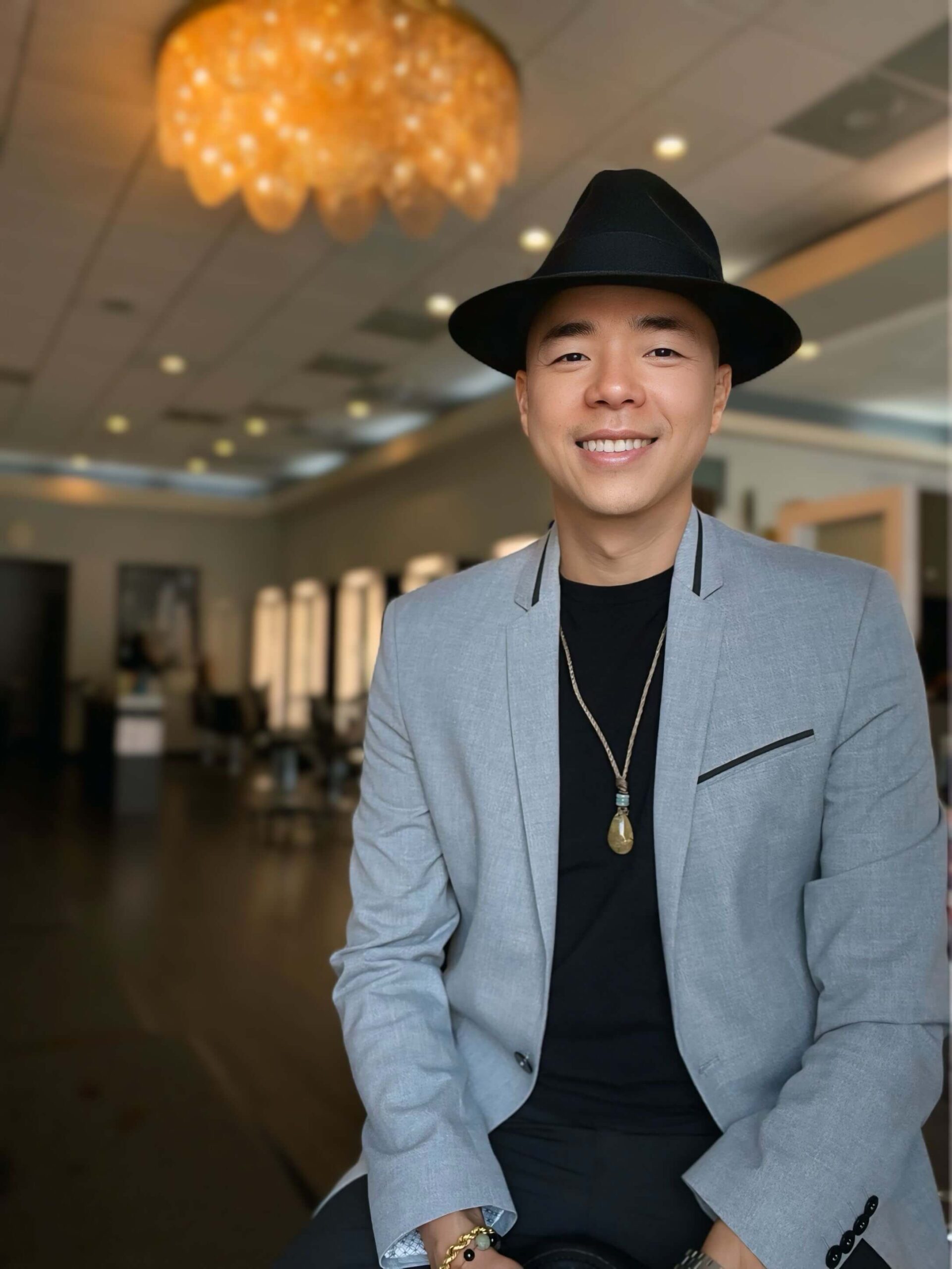 A person in a gray blazer and black hat smiling, standing in a modern interior with a chandelier.