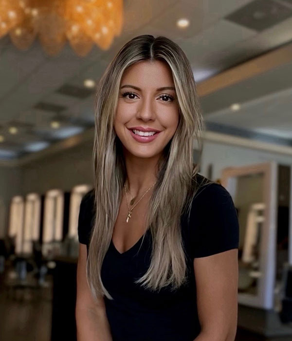 A woman with long, blonde hair and subtle balayage highlights smiles in a black top while standing indoors, surrounded by salon chairs and mirrors.