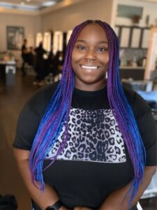 Daniella Coca beams, her long purple and blue braided hair cascading down as she rocks a black shirt adorned with a leopard print design. | Josephine's Salons and Spas