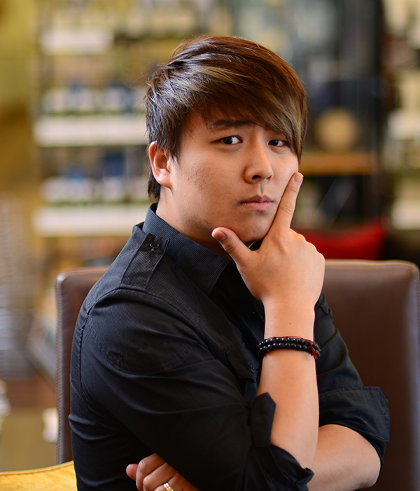 Person in a black shirt rests chin on hand, posing thoughtfully indoors with blurred background, showcasing his fresh men's cut.