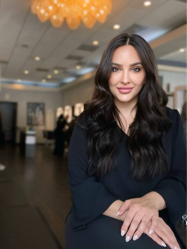 A woman with long dark hair sits in a modern, well-lit room, wearing a black top and smiling softly at the camera.