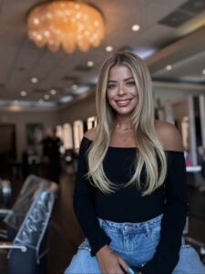 Patricia Cantrell, with her long blonde hair, sits smiling in a well-lit salon, radiating elegance in a black off-shoulder top and jeans. A chandelier sparkles overhead while mirrors reflect her joy. | Josephine's Salons and Spas