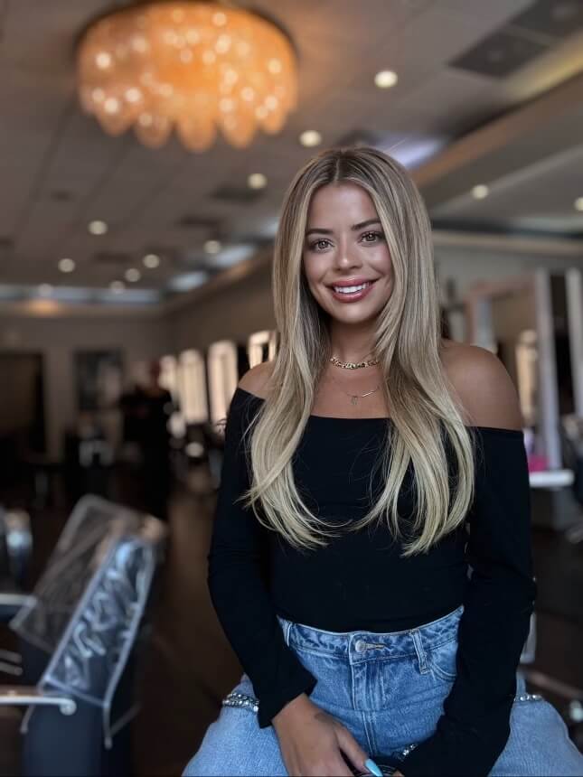 Person with long hair smiles in a salon, wearing a black top and blue jeans. A chandelier is visible in the background.