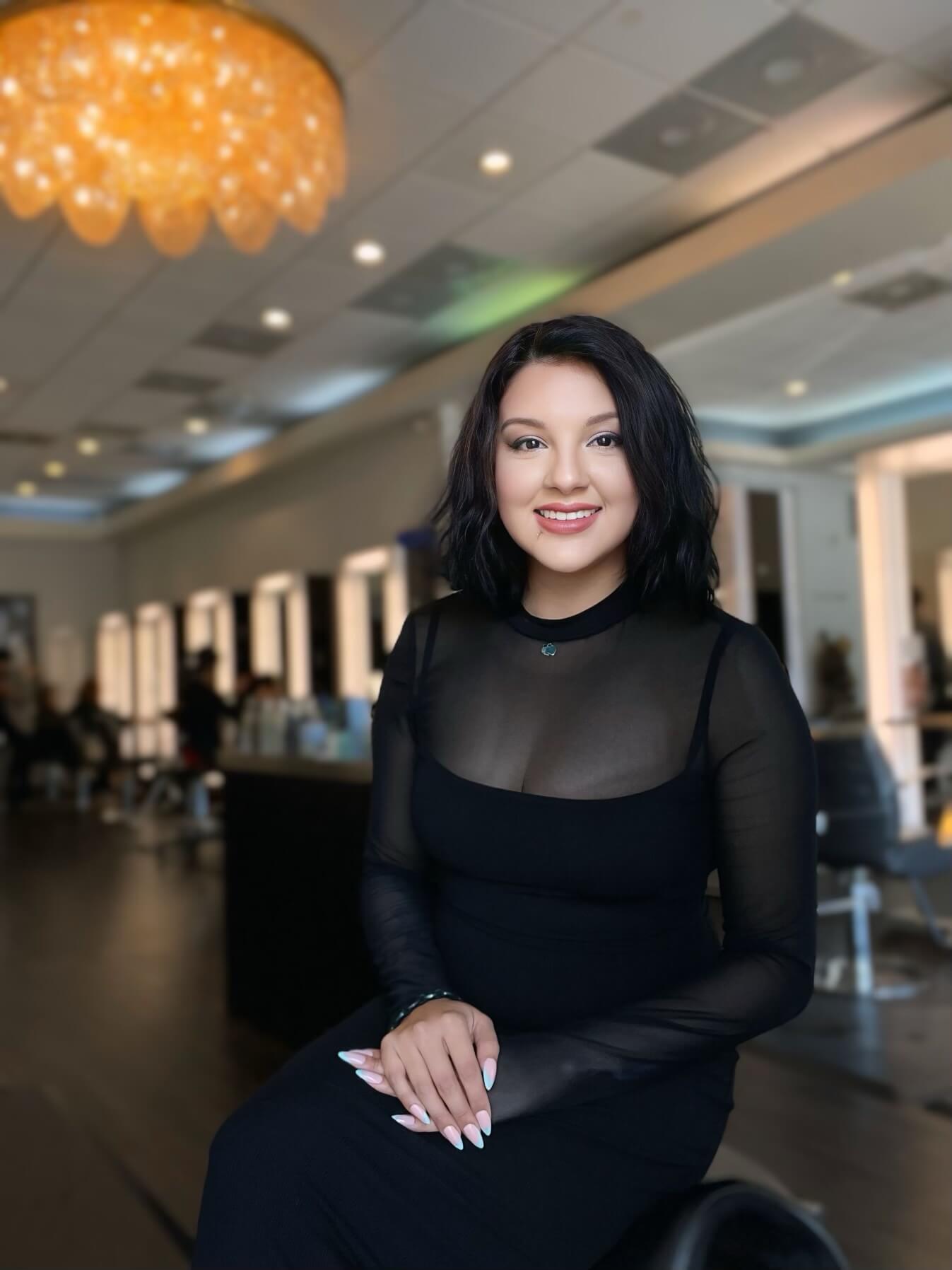 A woman in a black dress sits in a salon, smiling at the camera. The background shows salon chairs and mirrors, with warm lighting from a chandelier.