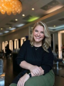 Kristy Monnin, with her blonde hair and black shirt, smiles in a well-lit room adorned with mirrors and hanging lights. | Josephine's Salons and Spas
