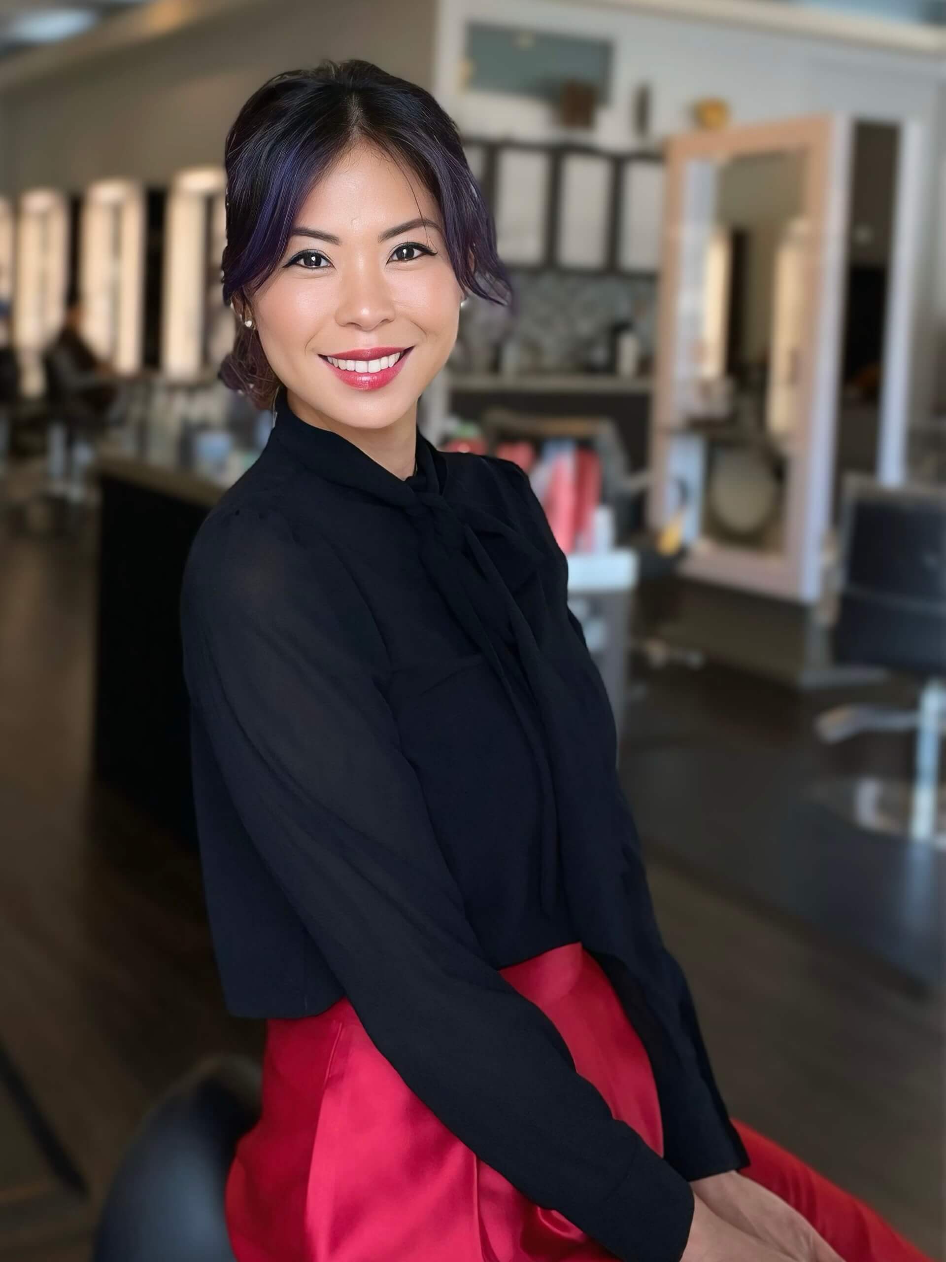 Person smiling in a salon, wearing a black top and red skirt. Background features salon mirrors and chairs.