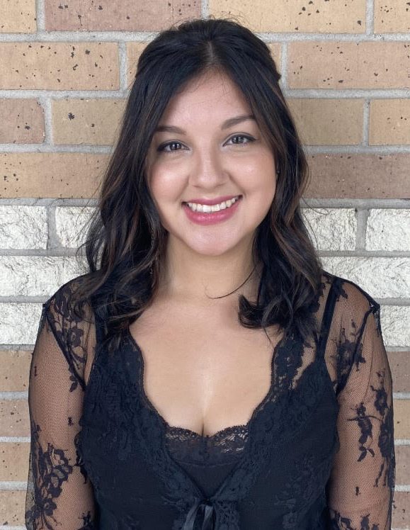 A woman with long dark hair and a black lace top smiles in front of a brick wall, exuding the serene confidence often seen after a visit to Josephine's Day Spa.