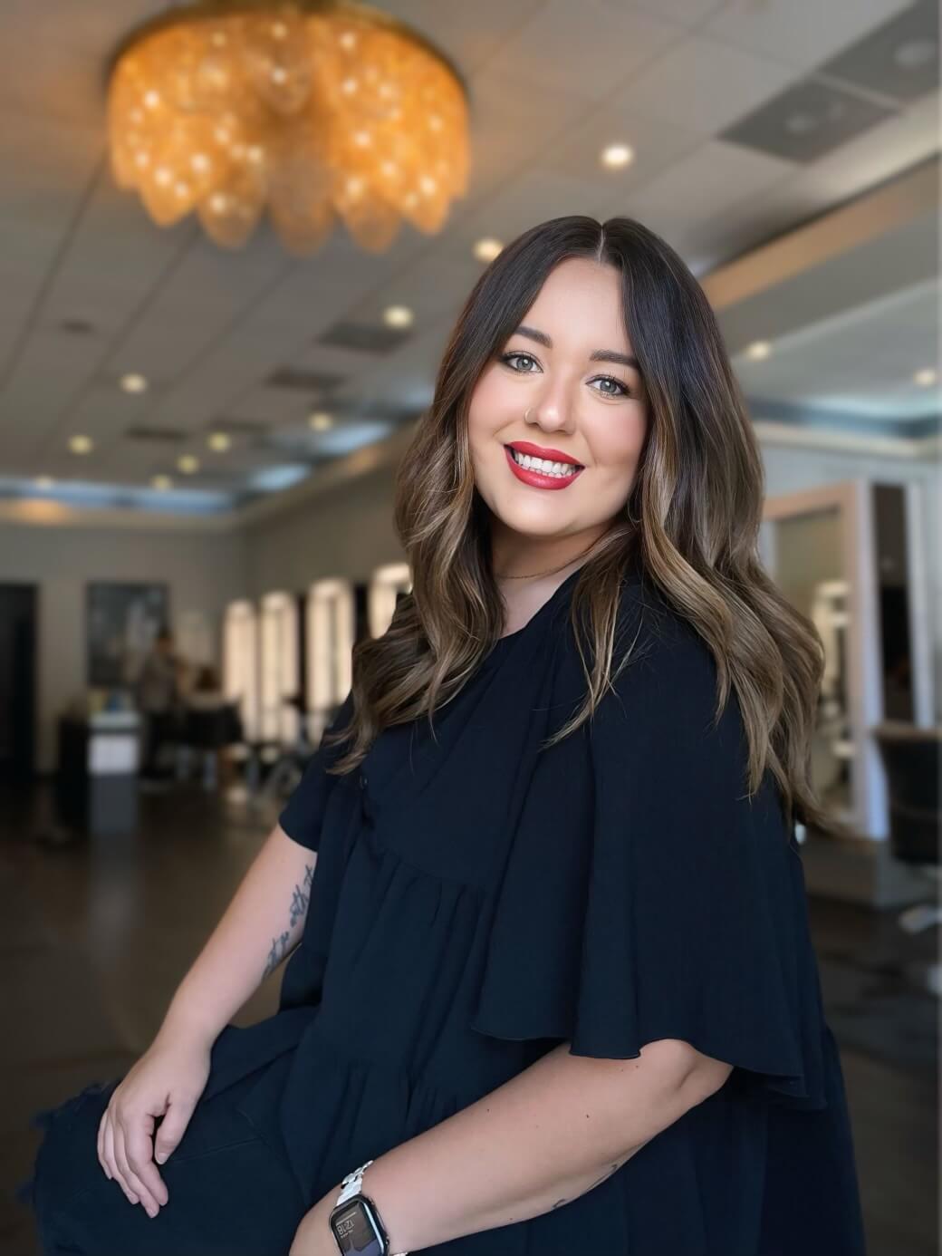 Woman with long brown hair and red lipstick smiles in a salon, sitting on a chair. A chandelier hangs from the ceiling.