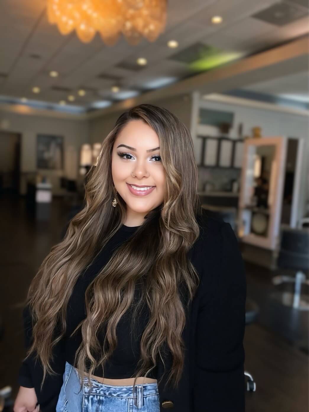 A person with long wavy hair smiles in a brightly lit room with salon chairs and mirrors in the background.