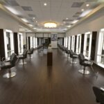 eldridge-11 Modern salon interior with rows of styling stations and mirrors on both sides, dark wood flooring, and a ceiling light fixture in the center. | Josephine's Salons and Spas