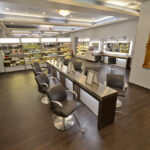 eldridge-5 A modern salon interior with rows of styling chairs and mirrors, sleek shelves lined with products, and a large gold-framed mirror on the wall. The floor is dark wood. | Josephine's Salons and Spas
