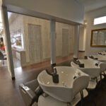 eldridge-6 A modern salon interior with a row of white shampoo sinks, styling chairs, a wall with bead curtain, and a large mirror. Bright lighting with wooden flooring. | Josephine's Salons and Spas