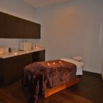 eldridge-9 A dimly lit spa room with a massage table covered in a brown blanket and white towel. Candles are placed on the table, and a counter with cabinets is against the wall. | Josephine's Salons and Spas