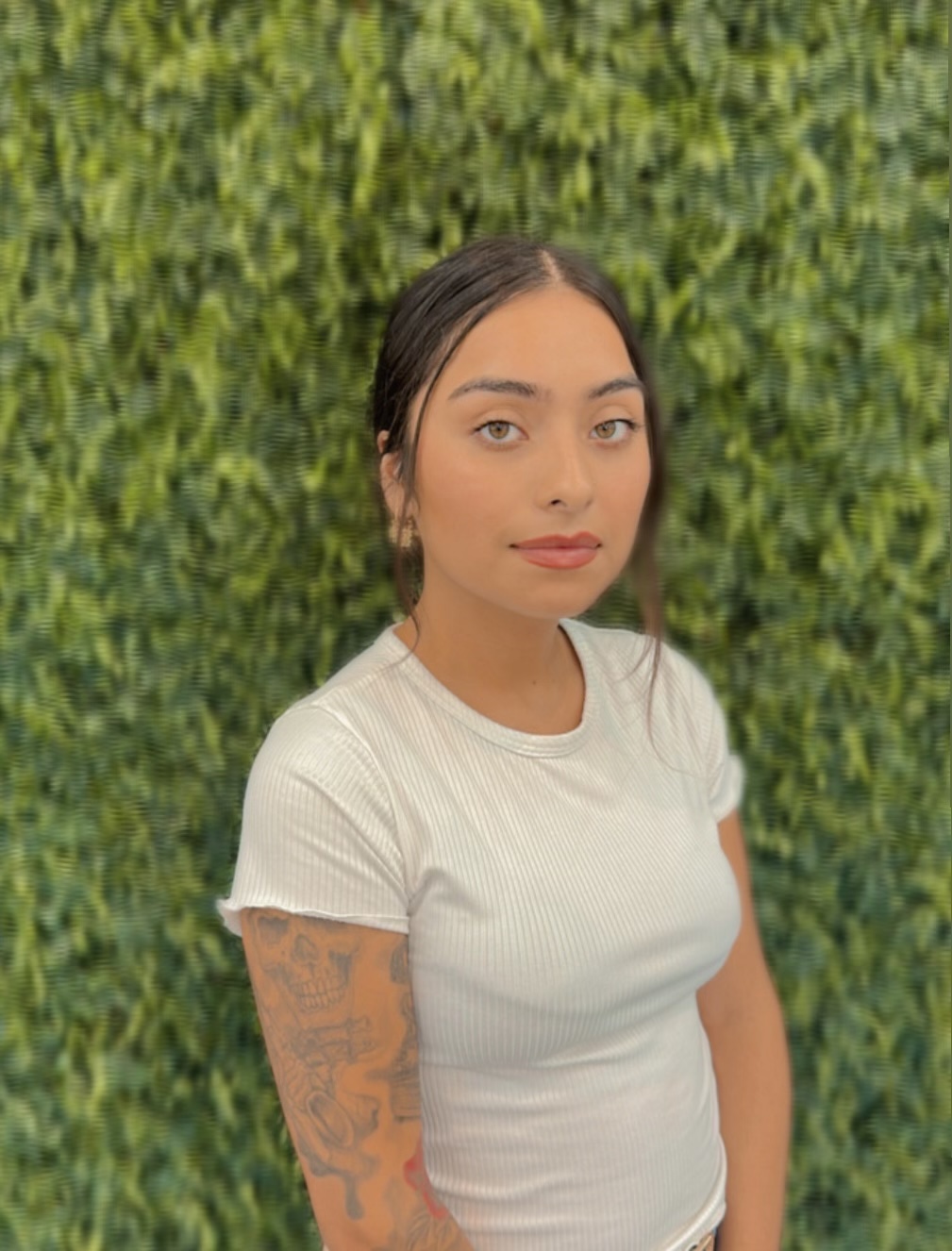 A woman with long dark hair and a tattoo on her left arm stands against a leafy green background, wearing a white ribbed short-sleeve shirt that perfectly complements the serene atmosphere of Josephine's Day Spa.