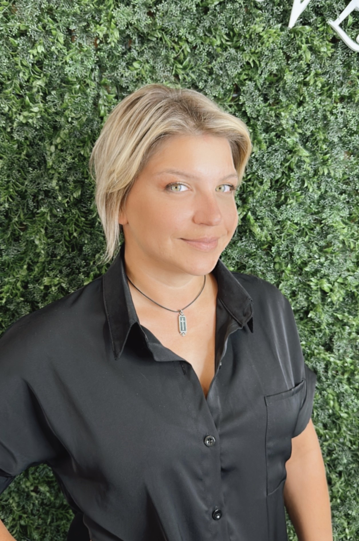 Person wearing a black shirt and necklace with a small pendant, standing against a green leafy background, smiling slightly at the camera, after visiting Josephine's Day Spa.