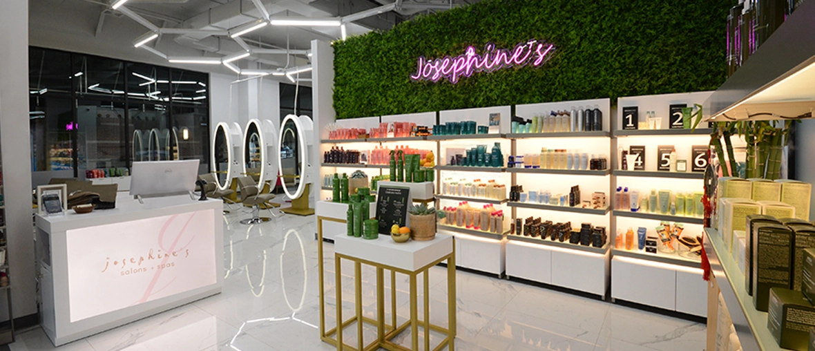 A salon interior with modern decor. Neon sign "Josephine's Day Spa", a reception desk, product display shelves, plants, and a seating area with illuminated circular mirrors are visible.