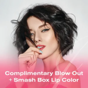 Woman with short brown hair wearing makeup, holding hair near mouth. Text overlay: "Complimentary Blow Out + Smash Box Lip Color—perfect for showcasing your balayage and highlights.