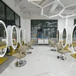 rice-village-2 A modern salon in Rice Village, Houston, boasts five styling stations with round mirrors, beige chairs, and geometric ceiling lights on a glossy floor. | Josephine's Salons and Spas