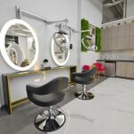 rice-village-7 Modern hair salon interior in Rice Village, featuring two styling chairs, illuminated mirrors, overhead hair dryers, and a small kitchenette. Gray flooring and wall-mounted foliage decor enhance the contemporary feel, making it a perfect stop during your shopping or dining outing. | Josephine's Salons and Spas