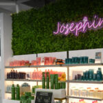 25-0190-InteriorHeader-P4-RiceVillage A modern salon interior with illuminated mirrors, a wall of hair products, and a neon sign reading "Josephine's" above a greenery-covered wall. | Josephine's Salons and Spas