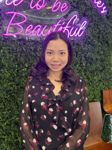 At our luxury Aveda salon and spa in Houston, TX, Tammy Nguyen shines with lush black hair and chic style beside a vibrant green wall and glowing "Beautiful" neon sign. | Josephine's Salons and Spas | Houston, TX