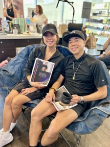 Two guests with stunning hair, dressed in black at Josephine’s Aveda Salon & Spa Houston, enjoy coffee and books in a stylish, cozy setting surrounded by luxury salon products. | Josephine's Salons and Spas | Houston, TX