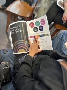 Guest at Josephine’s Luxury Aveda Salon & Spa in Houston holds booklet on “Behaviors That Drive Benchmarks,” showing commitment to excellence; camera and phone on lap. | Josephine's Salons and Spas | Houston, TX