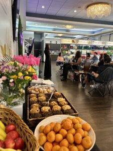 Inside Josephine’s luxury Aveda salon in Houston, a woman speaks to guests. Muffins, fruit, and flowers are displayed by shelves of premium hair care products in a stylish spa setting. | Josephine's Salons and Spas | Houston, TX